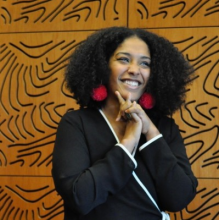 Suhaly Bautista-Carolina headshot: a caramel-skin toned woman with black, curly hair stands smiling against a brown, wooden wall with swirling grooves. She is wearing a black jumpsuit with white stripes and puffy pink earrings. Her hands are clasped together against her chin.