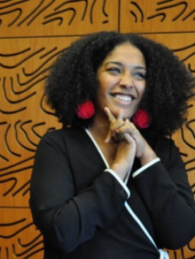 Suhaly Bautista-Carolina headshot: a caramel-skin toned woman with black, curly hair stands smiling against a brown, wooden wall with swirling grooves. She is wearing a black jumpsuit with white stripes and puffy pink earrings. Her hands are clasped together against her chin.